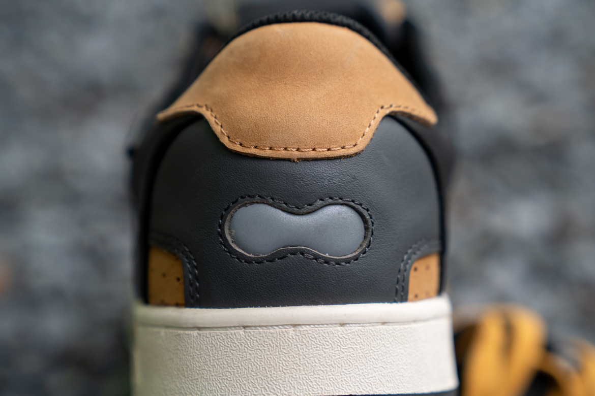 Close-up view of the back of a sneaker, featuring a combination of smooth black leather and tan suede, with a gray emblem in an oval shape and stitching details. The sole of the shoe is white with a textured finish. The background is blurred, emphasizing the shoe design.