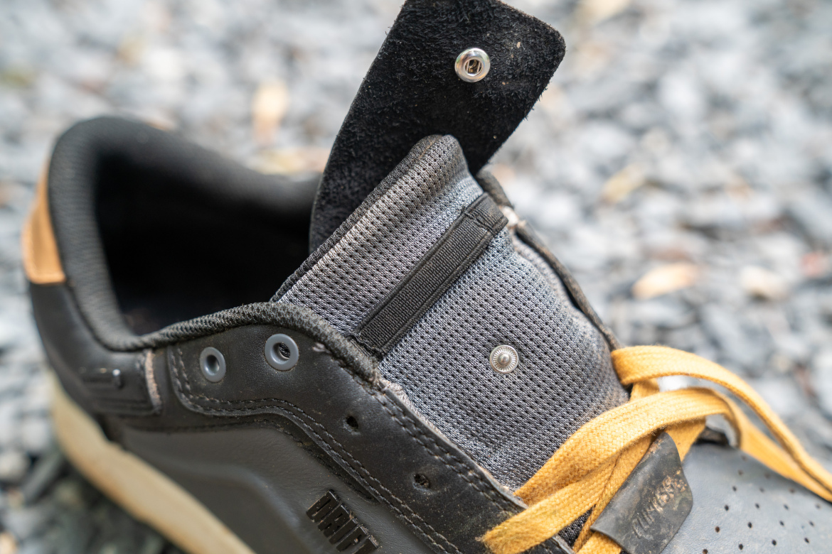 Close-up view of the tongue and lacing area of a black sneaker with yellow laces. The sneaker features a mesh tongue and a gray fabric overlay, with metal eyelets and a snap button at the top. The background shows a textured, gravel surface.
