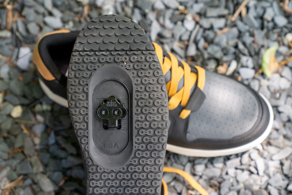 Close-up view of the sole of a black and gray sneaker with yellow laces, featuring a clip for attaching to a cycling pedal, resting on a bed of small gray stones.