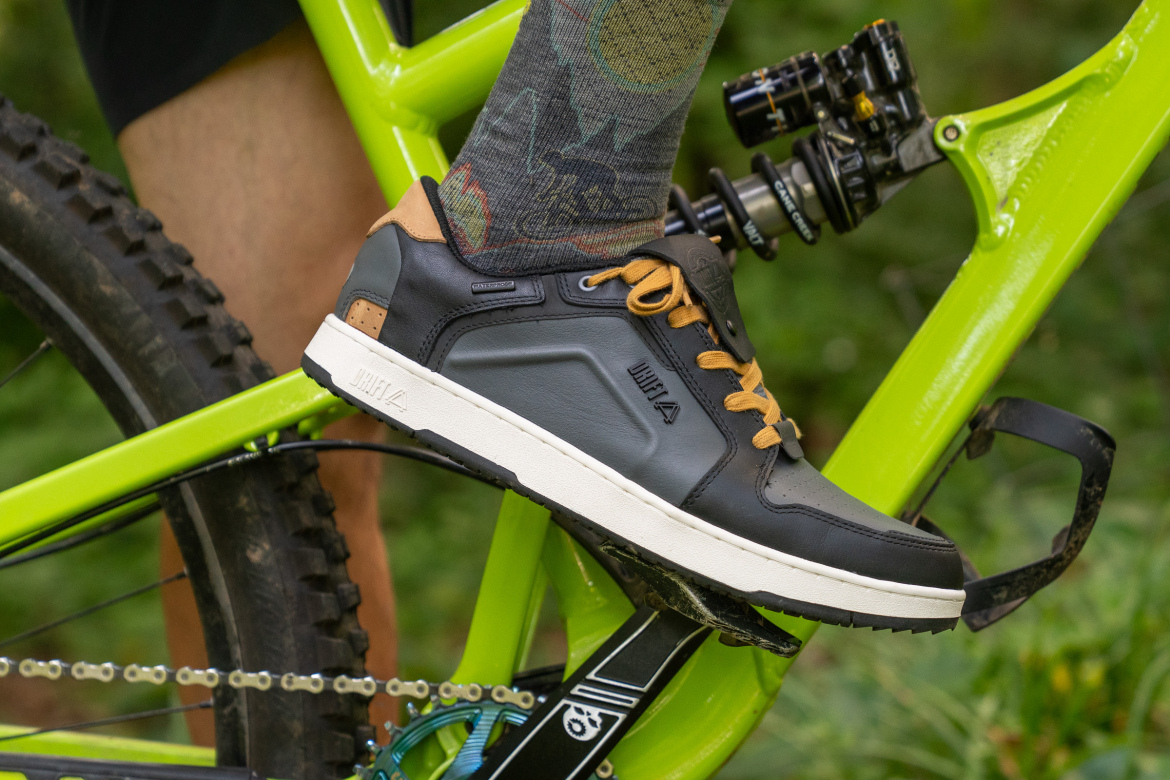 Close-up of a cyclist's foot wearing a black and brown biking shoe, placed on a pedal of a bright green mountain bike. The cyclist is also wearing a patterned sock, and the bike features elements of off-road design, showcasing a natural outdoor background.