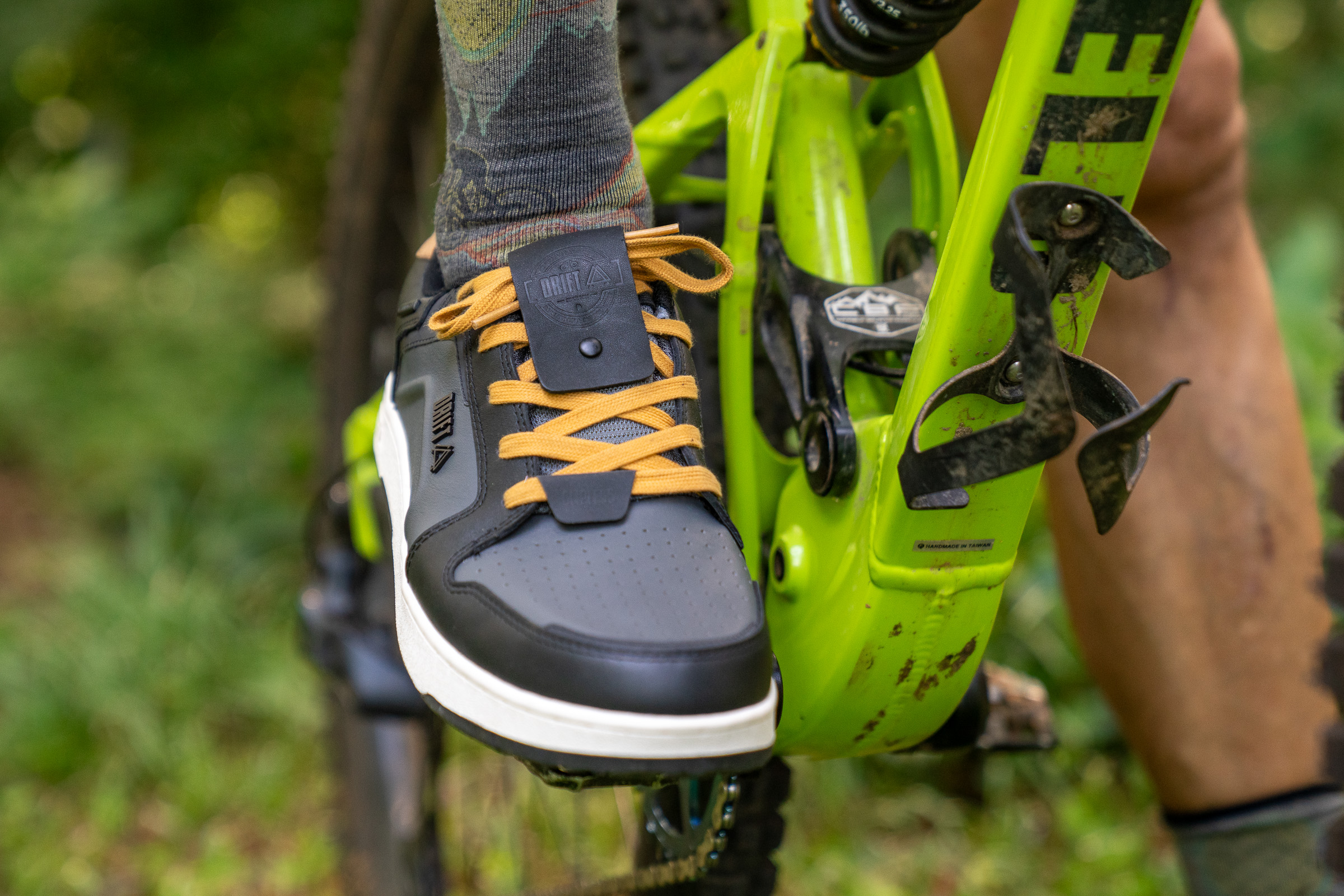 Close-up of a black and orange biking shoe resting on a green bicycle pedal, with a blurred background of foliage. The shoe features a unique design with perforated detailing and is paired with patterned socks. The bike frame and pedal are visible, showing some dirt, indicating outdoor use.