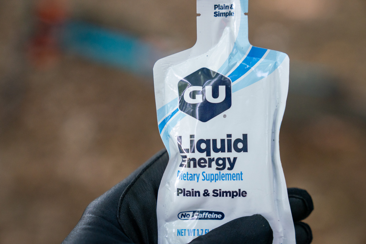 A close-up image of a GU Liquid Energy dietary supplement pouch, labeled "Plain & Simple," held in a gloved hand. The pouch design features a blue and white color scheme, indicating it's caffeine-free, with a net weight of 1.7 fluid ounces. The background is blurred, suggesting an outdoor setting.