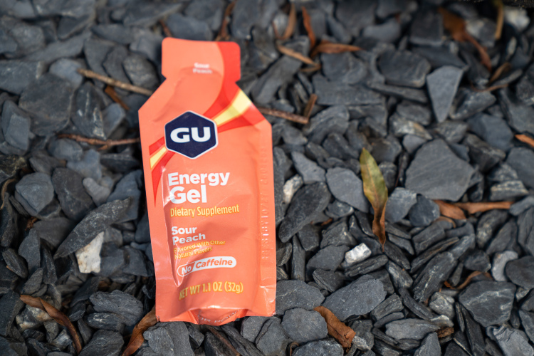 An energy gel packet on a bed of dark gravel stones. The packet is orange with a label that reads "GU Energy Gel," specifying the flavor as "Sour Peach" and noting it contains no caffeine.