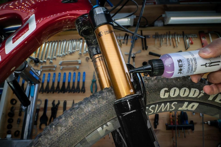 A close-up view of a mountain bike's front suspension fork being serviced. A person's hand is applying suspension oil from a bottle labeled "Joe's Suspension Fluid" into the fork, while an array of bicycle tools and parts is visible in the background. The bike has a maroon frame and a tire with the brand "Good Year."