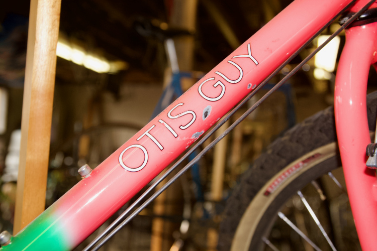 Close-up of a colorful bicycle frame showing the name "OTIS GUY" etched in white on a pink gradient surface, with visible wear and scratches, set in a cluttered background.