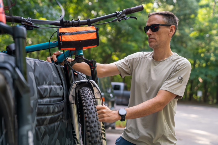 A man wearing sunglasses and a light-colored shirt secures a mountain bike onto a bike rack, surrounded by lush green trees in the background. He is focused on adjusting the bike, which has a bright orange bag attached to the handlebars.