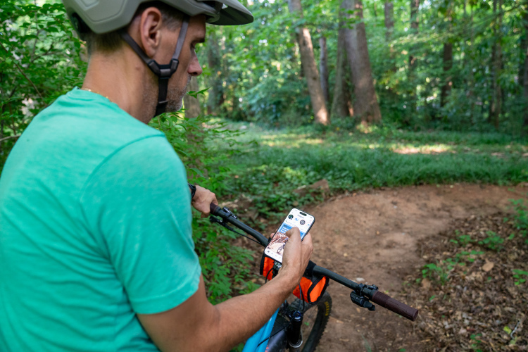 A person wearing a helmet sits on a mountain bike, holding a smartphone while surrounded by lush greenery on a dirt path in the woods.