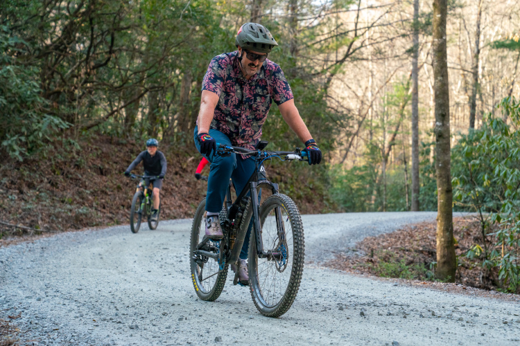 A man in a floral-patterned shirt and helmet rides a black mountain bike on a gravel winding path, surrounded by trees. In the background, a child on another bike follows closely behind. The scene captures an outdoor biking adventure in a wooded area.