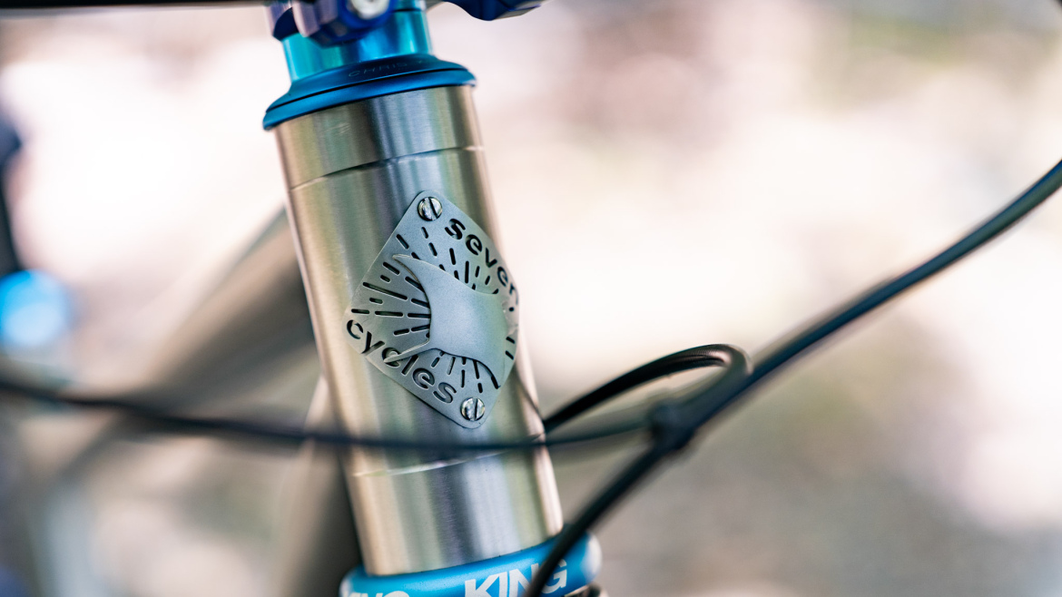 Close-up image of a bicycle's head tube featuring a distinctive metallic badge with the text "seven cycles," alongside cables connected to the frame, set against a blurred background.