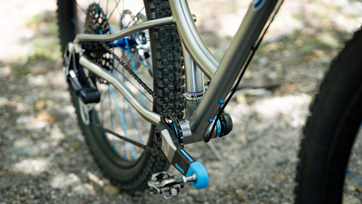 A close-up view of a bicycle's rear drivetrain, showcasing a silver frame alongside blue accents. The shot captures the chain, cog, and pedal area on a rugged surface, with blurred greenery in the background.