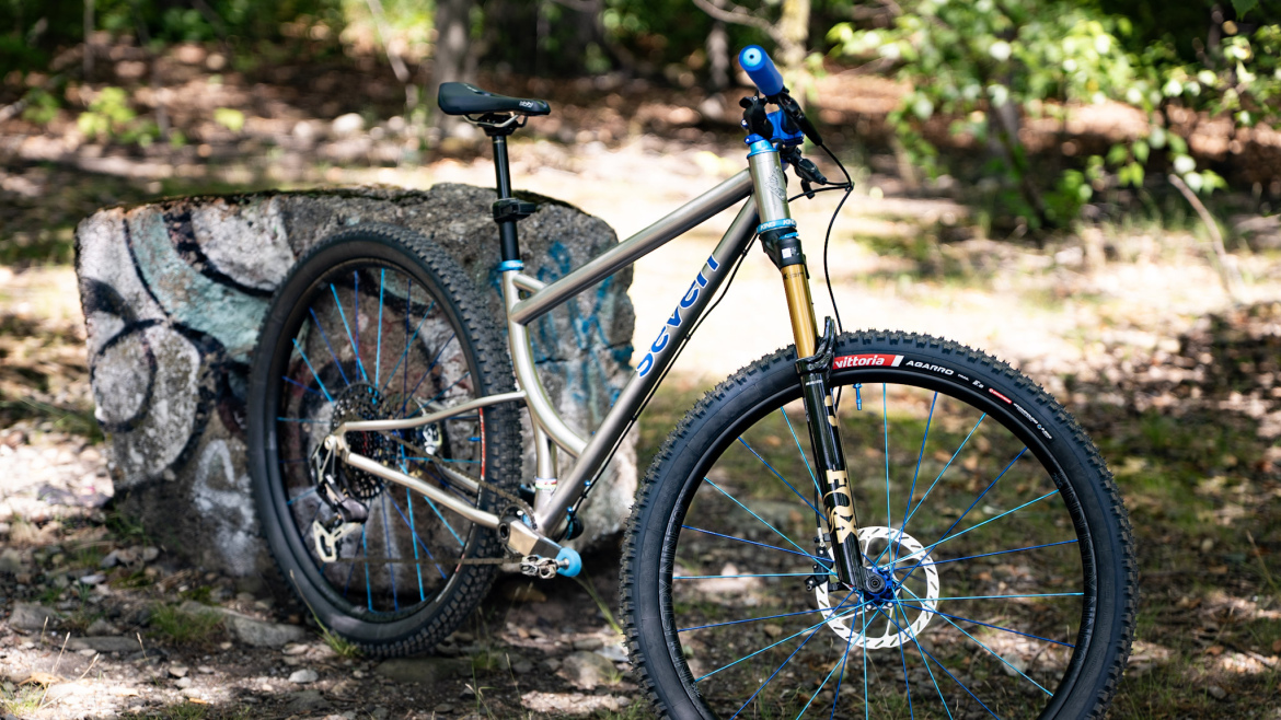 A high-quality mountain bike is positioned next to a textured stone with graffiti in a wooded outdoor setting. The bike features a sleek silver frame with blue accents, thick tires designed for off-road terrain, and a sturdy front suspension fork, suggesting it's built for trail riding. Sunlight filters through the trees, highlighting the bike and the natural surroundings.
