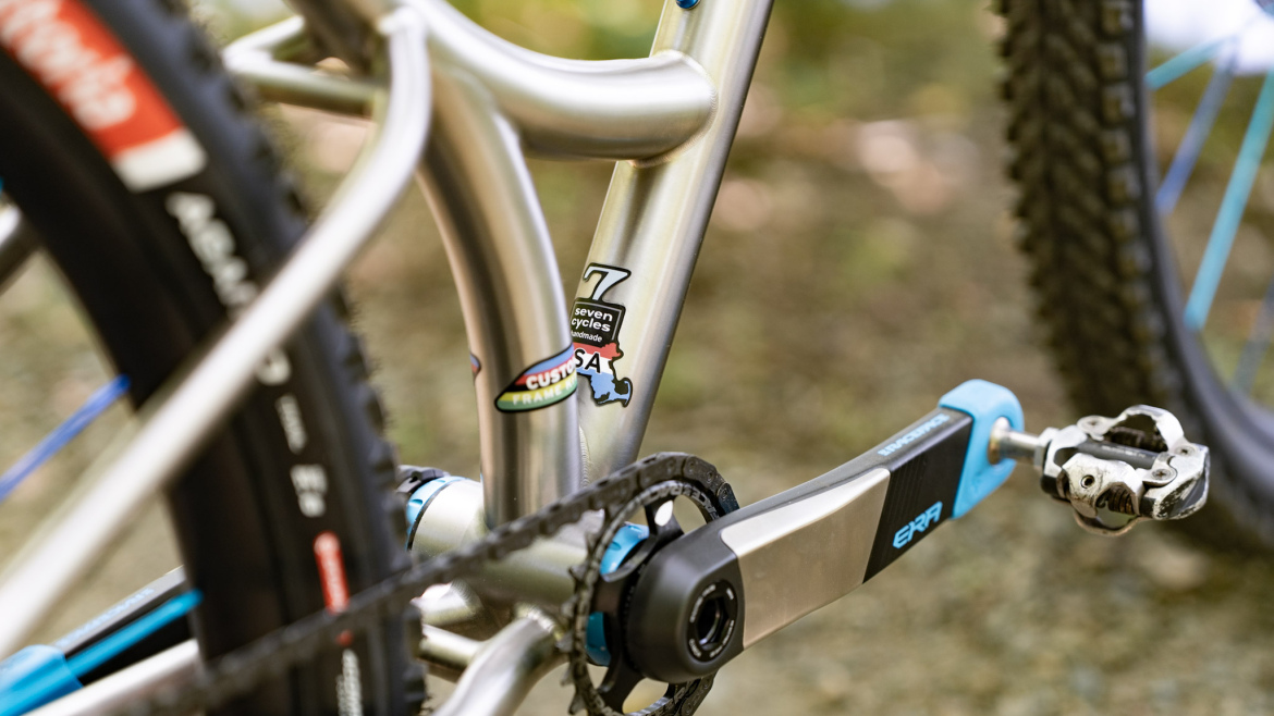 A close-up view of a bicycle frame showcasing a silver metal construction, featuring a sticker that reads "seven cycles" and another that says "custom." The image captures part of the crankset and pedal, along with a glimpse of the black tire with blue detailing. The background is blurred, highlighting the bike's components.