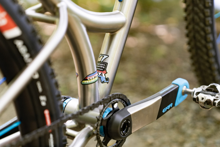 A close-up view of a bicycle frame showcasing a silver metal construction, featuring a sticker that reads "seven cycles" and another that says "custom." The image captures part of the crankset and pedal, along with a glimpse of the black tire with blue detailing. The background is blurred, highlighting the bike's components.