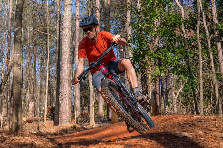 A mountain biker in an orange shirt and gray shorts navigates a dirt trail in a wooded area, leaning into a turn while riding a black mountain bike with thick tires. Trees and greenery surround the trail, showcasing a sunny day in nature.