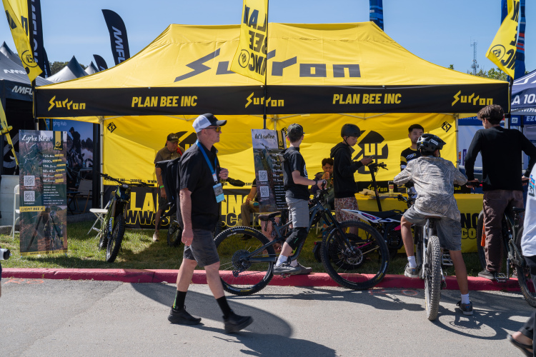 A busy outdoor event featuring a yellow tent with Surron branding, where visitors are engaging with staff and exploring mountain bikes. Several people, including children and adults, are gathered around the bikes, while one man walks by in a black shirt and cap. Prominent banners and promotional materials are displayed nearby, highlighting the products and services offered.