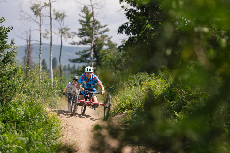 Two individuals riding mountain bikes along a dirt path surrounded by greenery. One rider is in the foreground, wearing a blue shirt and helmet, while the second rider is further back. The scene captures a sunny day with trees and mountains in the background.