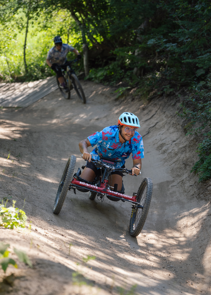 A person riding a handcycle down a dirt trail, smiling and wearing a colorful shirt and a helmet. In the background, another cyclist can be seen navigating the same path. The scene is surrounded by green trees and sunlight filtering through the leaves, creating a lively and adventurous atmosphere.