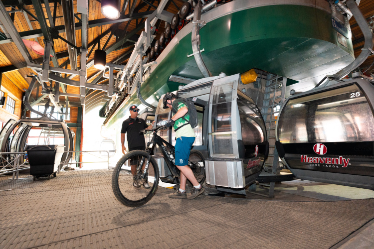 A man in a helmet and riding gear is preparing to board a gondola with his mountain bike in a lift station. A staff member stands nearby, providing assistance. The interior features wooden beams and several gondola cars, with a large gondola above.