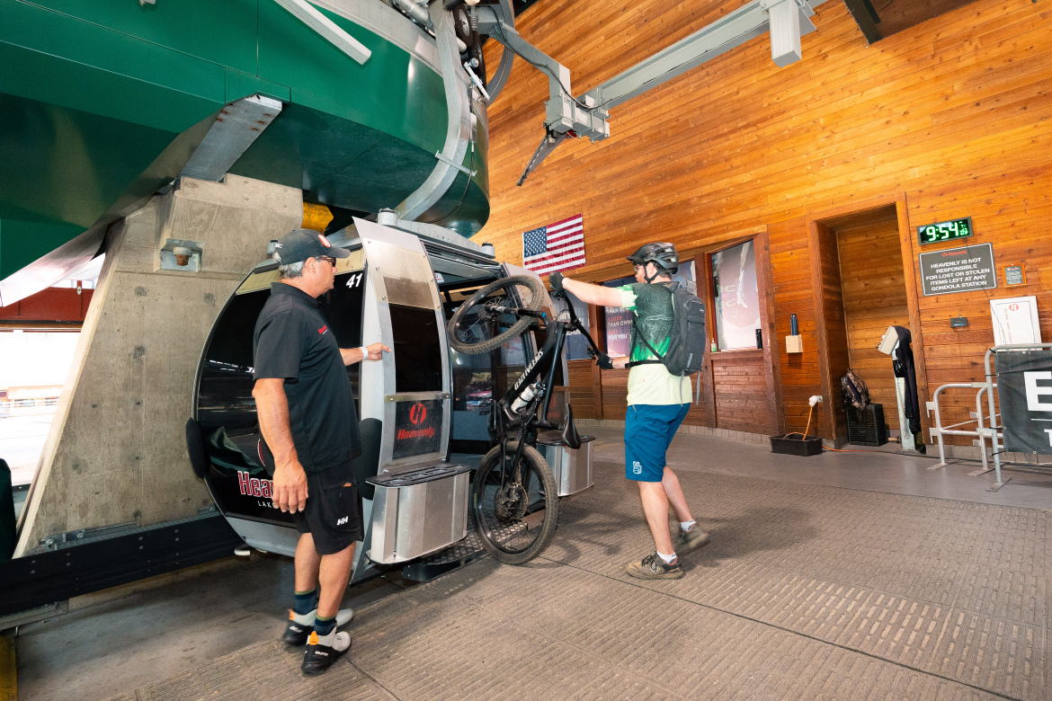 A man in a black shirt assists a cyclist with a mountain bike as he prepares to load it into a gondola at a lift station. The interior features wooden walls and a clock displaying 9:54. An American flag is visible in the background.