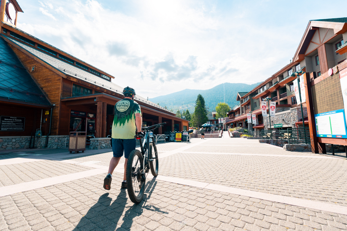 A cyclist wearing a helmet and a colorful shirt walks a mountain bike through a pedestrian area lined with shops and buildings. In the background, mountains rise under a partly cloudy sky. The scene captures a vibrant, outdoor atmosphere in a recreational area.