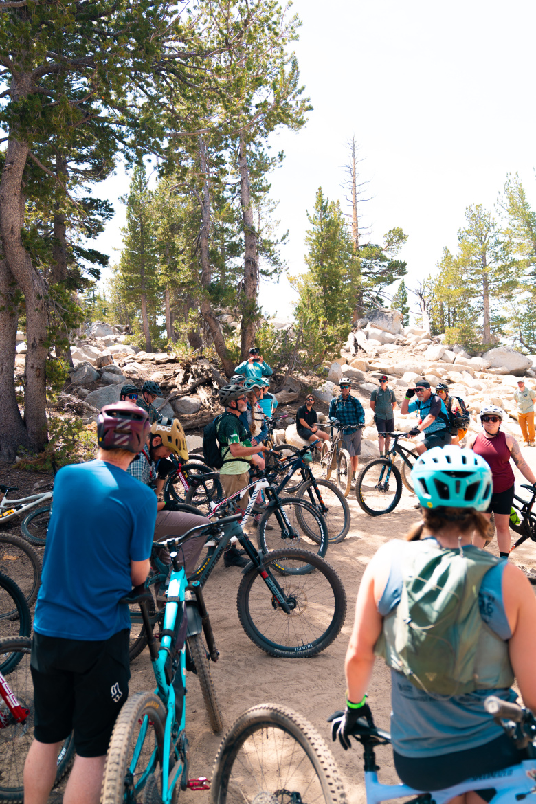A group of mountain bikers gathered on a dirt path surrounded by trees and rocky terrain, discussing a ride. Several bicycles lean against each other while participants wear helmets and cycling gear, showing a range of styles and colors. The setting is bright and sunny, indicating a pleasant outdoor atmosphere.
