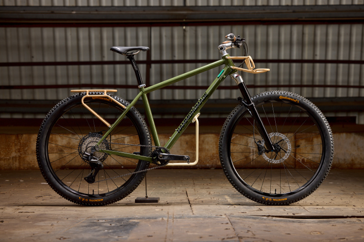 A green mountain bike with an alloy gold rear rack, mounted on a wooden floor in an industrial setting with a corrugated metal background. The bike features black tires with orange lettering, a black fork, and a clean, minimalist design.