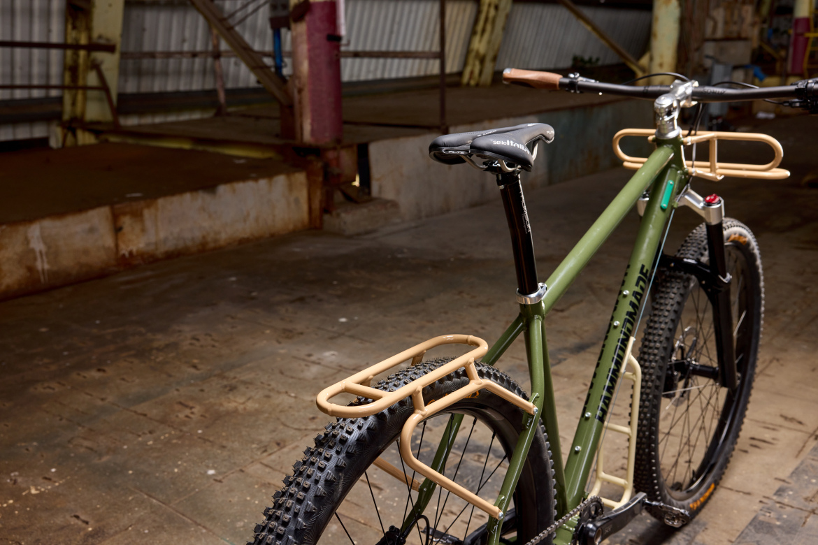 A close-up view of a green bicycle with a minimalist design, featuring a matte finish and leather grips. The bike has a prominent rear rack in a contrasting tan color, mounted on a thick tire suitable for off-road use. The background shows a rustic indoor setting with metal beams and a weathered floor, adding an industrial aesthetic to the scene.