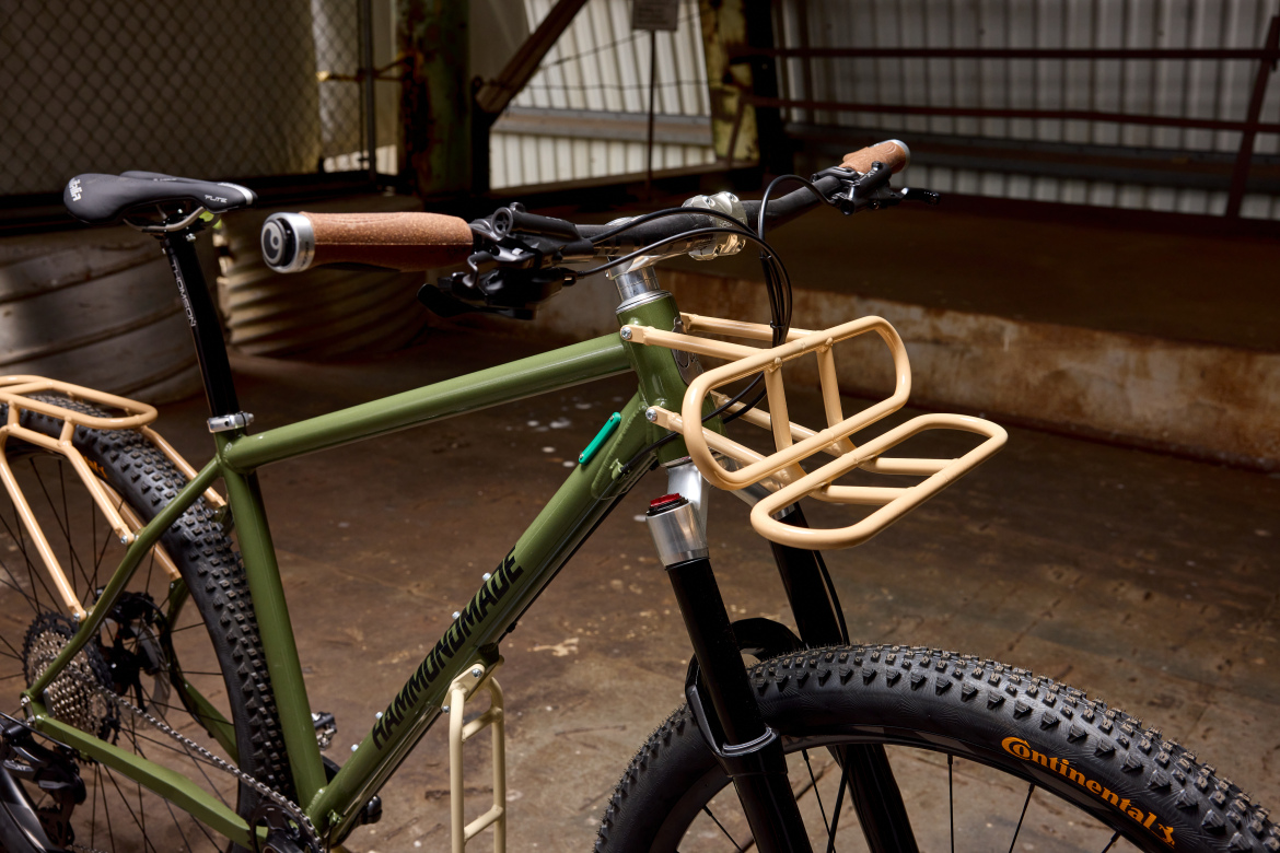 A close-up view of a green bicycle featuring a front rack. The bike has a black front fork, chunky tires with prominent treads, and cork handle grips. The background includes a textured floor and industrial setting with a fence and overhead structure.