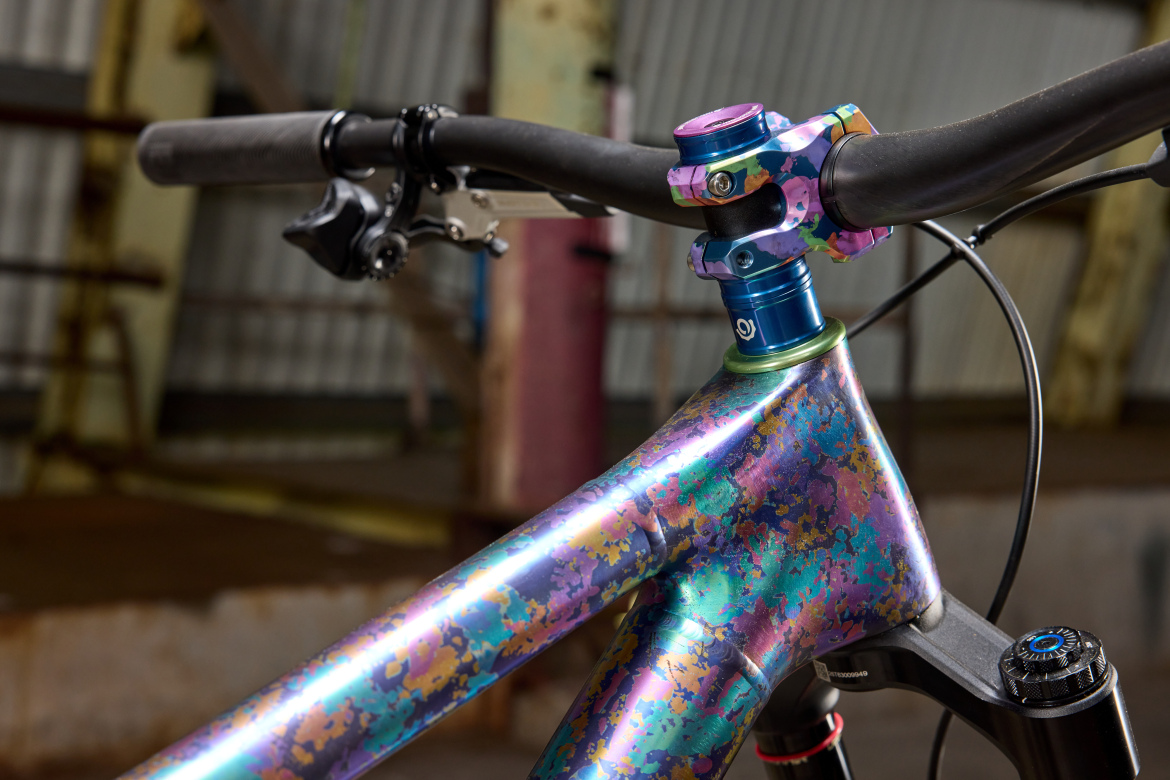 Close-up view of a colorful mountain bike's handlebars and stem, featuring an iridescent frame with a vibrant pattern and a blue and pink stem. The background shows an industrial setting with aged metal and beams.