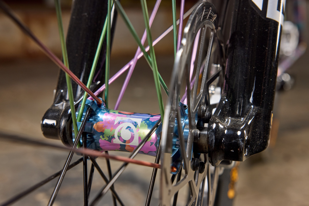 Close-up view of a bicycle hub showing colorful spokes, with a vibrant, multicolored design on the hub itself. The background is slightly blurred, highlighting the intricate details of the wheel and spokes.