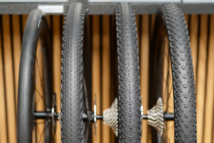 Close-up view of three bicycle tires mounted on a rack, showcasing different tread patterns. The tires are black with various textures, positioned in front of a blurred background of wooden slats. The image captures the details of the tires' designs.