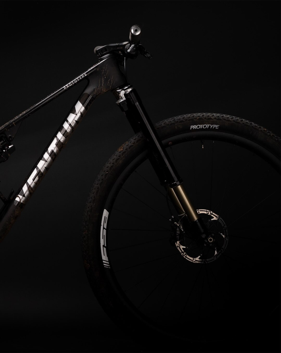 Close-up view of a mountain bike with a black and gold color scheme, featuring a dirty frame and tires. The bike's front fork is prominent, showcasing its suspension components, while the wheel is partially visible, highlighting the tread and brake disc. The background is solid black, emphasizing the bike's design and features.