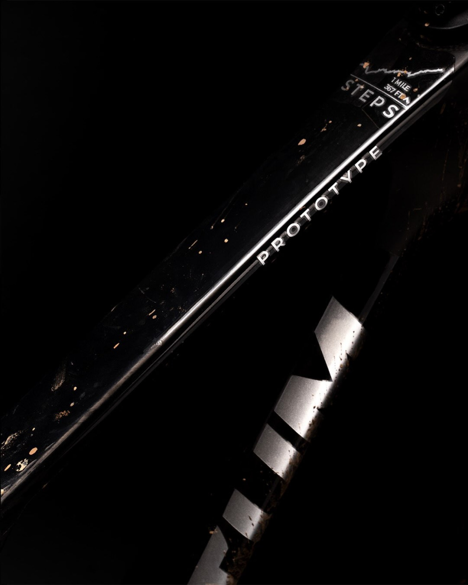 Close-up view of a black bicycle frame with the word "PROTOTYPE" and "STEPS" visible, set against a dark background. The frame shows signs of dirt and wear, indicating usage.
