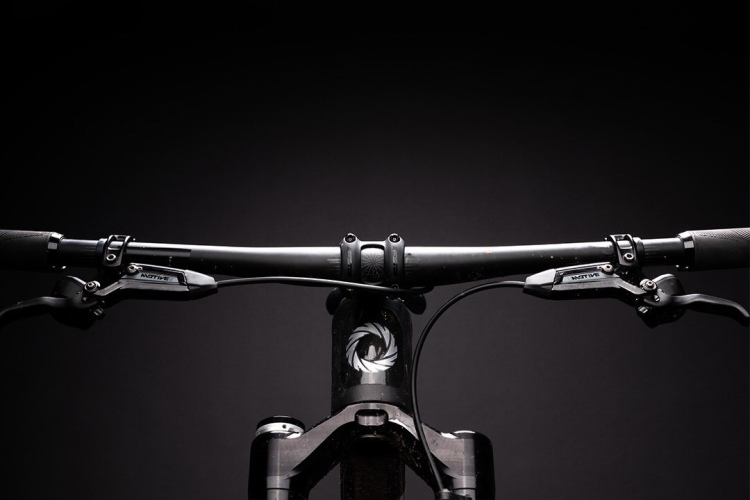 Close-up view of a mountain bike's handlebars and front wheel, set against a dark background with a bright overhead light illuminating the scene. The image highlights the bike's frame details, tires, and control components, emphasizing a clean and modern design.