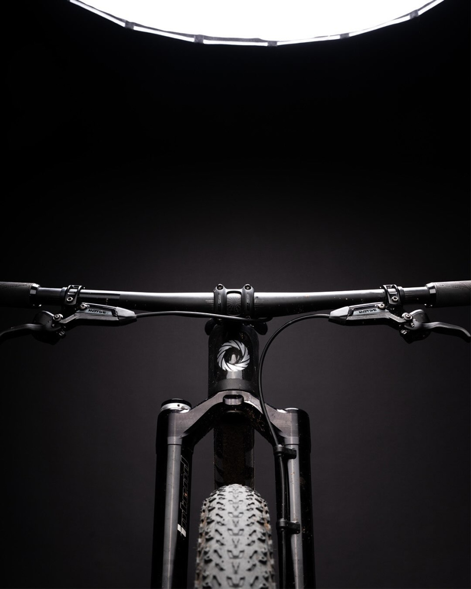 Close-up view of a mountain bike's handlebars and front wheel, set against a dark background with a bright overhead light illuminating the scene. The image highlights the bike's frame details, tires, and control components, emphasizing a clean and modern design.