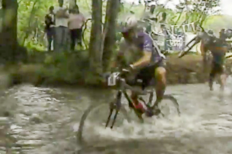 A mountain biker navigates through a stream during an off-road cycling event, with bystanders watching from the shoreline. The scene is surrounded by lush greenery, typical of a natural setting for outdoor sports.