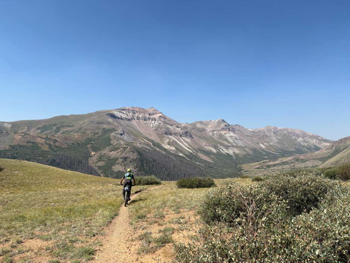 A person riding a mountain bike on a narrow dirt trail through a grassy landscape, with expansive mountains in the background under a clear blue sky.