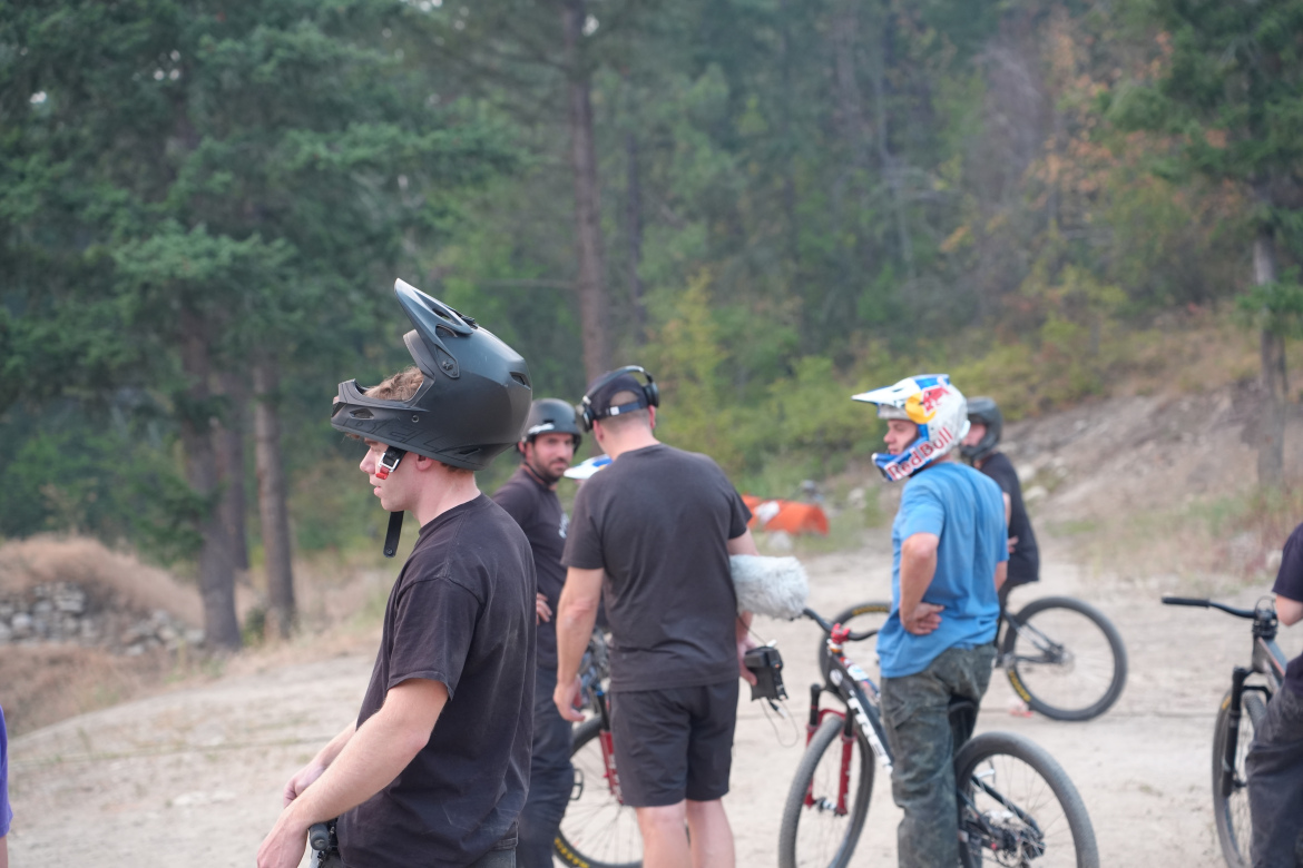 A group of mountain bikers wearing helmets in a wooded area, some holding bikes while conversing. The scene captures a casual gathering of riders preparing for a biking session.