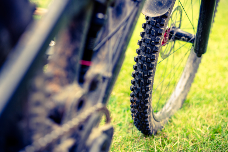 Close-up view of a mountain bike rear tire on grassy terrain. The tire features a textured tread pattern, and part of the bike's frame is visible in the background, showcasing dirt accumulation. The focus is on the tire, highlighting its rugged design suitable for off-road cycling.