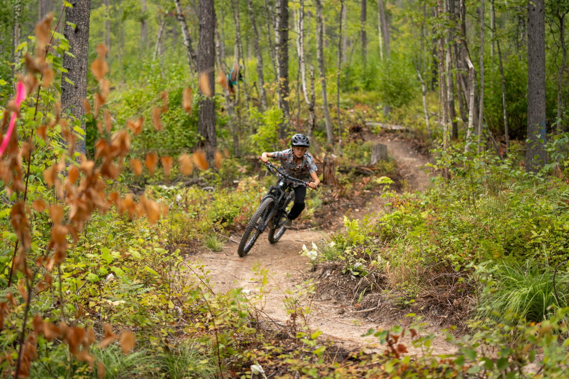 A young person riding a mountain bike on a forest trail, leaning into a turn. The scene is surrounded by greenery and trees, with some colorful markers indicating the biking path.