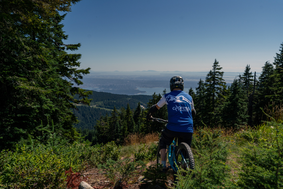 A mountain biker wearing a blue shirt and a helmet is sitting on a bike, overlooking a lush green landscape with trees and a distant view of a body of water and mountains under a clear blue sky.
