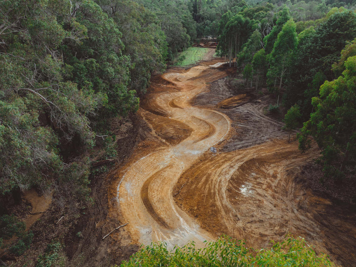 Aerial view of a winding dirt trail being constructed through a forested area, surrounded by lush green trees and earth that has been recently disturbed. The path features smooth curves and sections of exposed soil, indicating ongoing development in a natural setting.