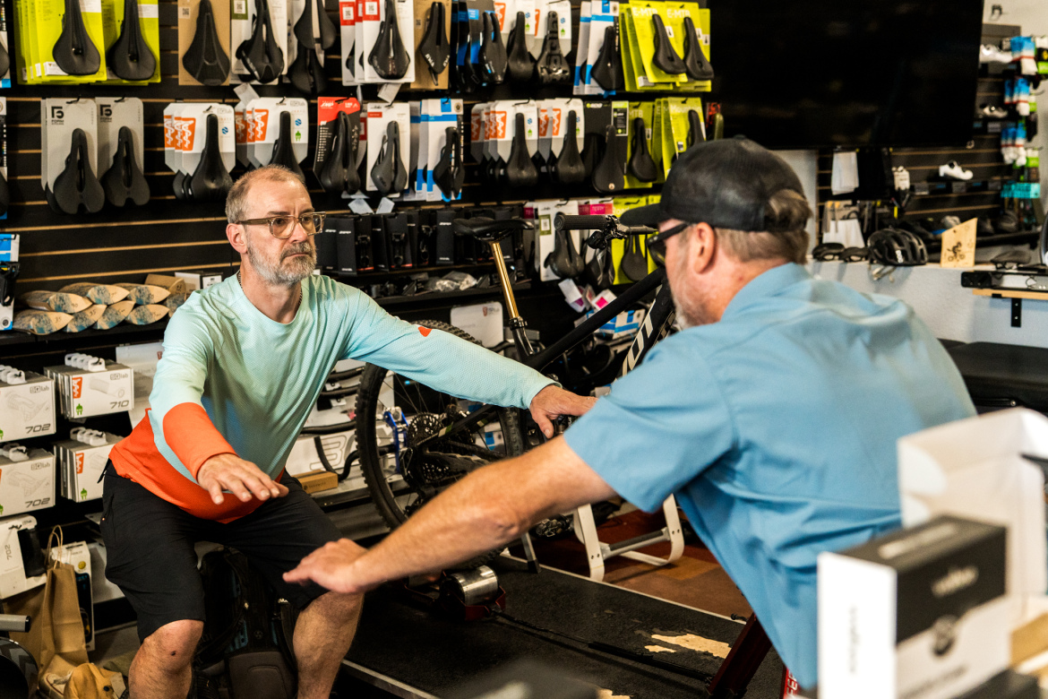 A man demonstrating a cycling position on a stationary bike, while another man, facing him, provides guidance. The store is well-stocked with various bicycle accessories and parts visible in the background.