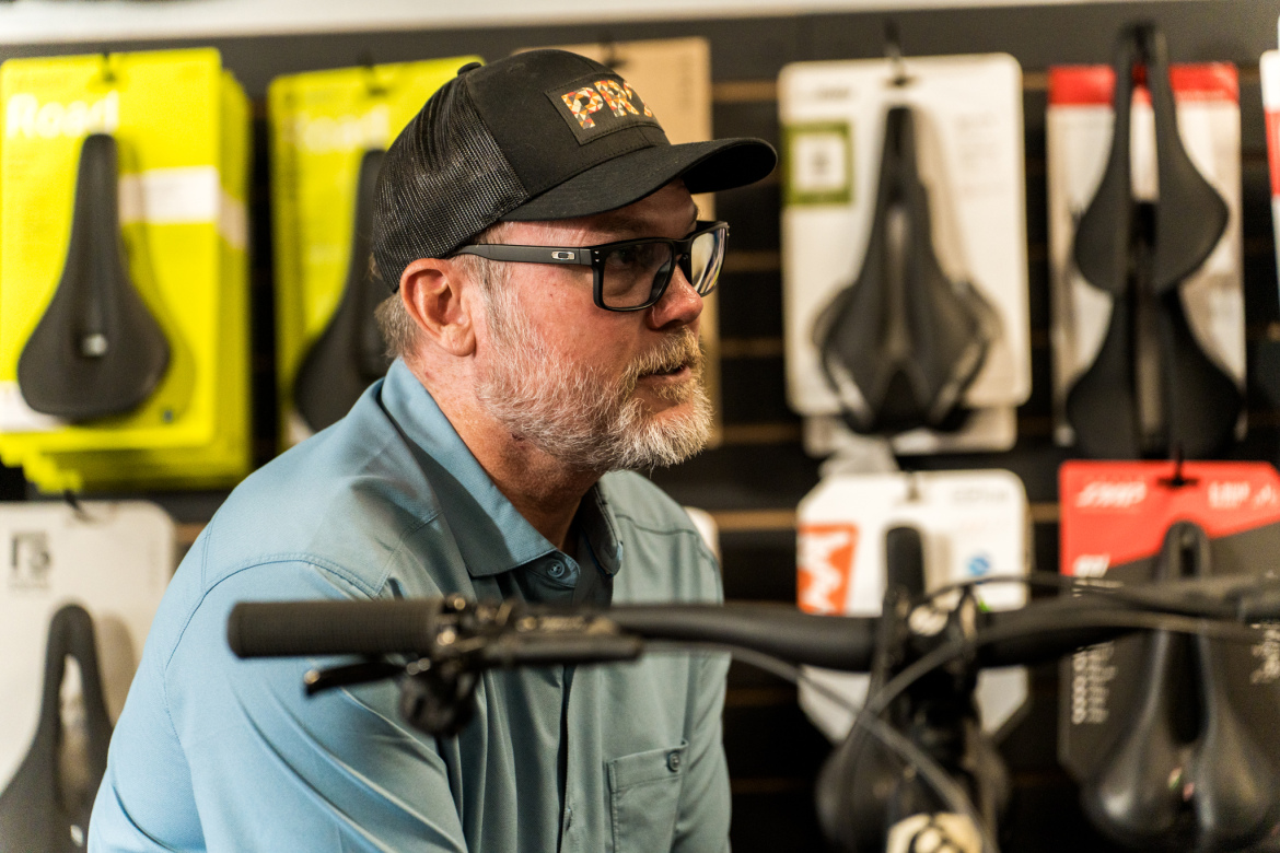 A middle-aged man with glasses and a baseball cap sits thoughtfully in a bike shop, surrounded by various bike seats displayed on the wall behind him. He is wearing a light blue shirt and is positioned next to a bicycle, with his hands resting on the handlebars.