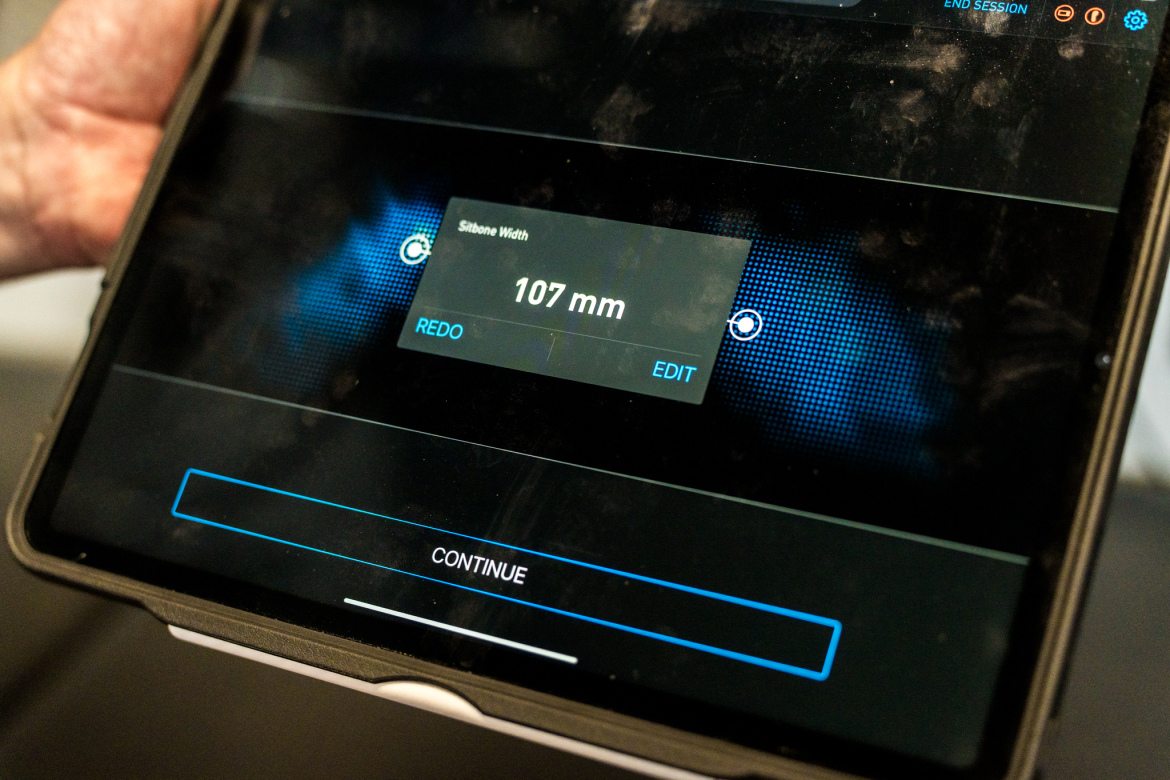 A close-up view of a tablet screen displaying a measurement of "107 mm" for "Sitbone Width," with options to "REDO" or "EDIT." The background features a blue dotted design, and there is a "CONTINUE" button at the bottom of the screen.