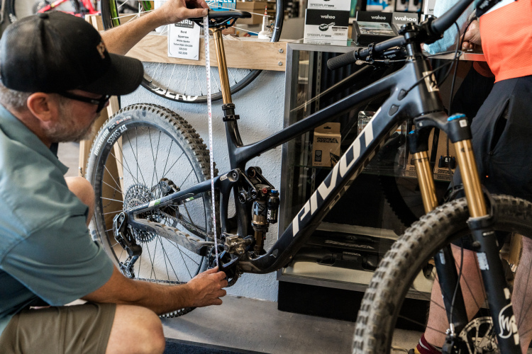 A person measuring the bottom bracket of a black Pivot mountain bike while another individual observes, inside a bike shop with various bicycle accessories displayed in the background.