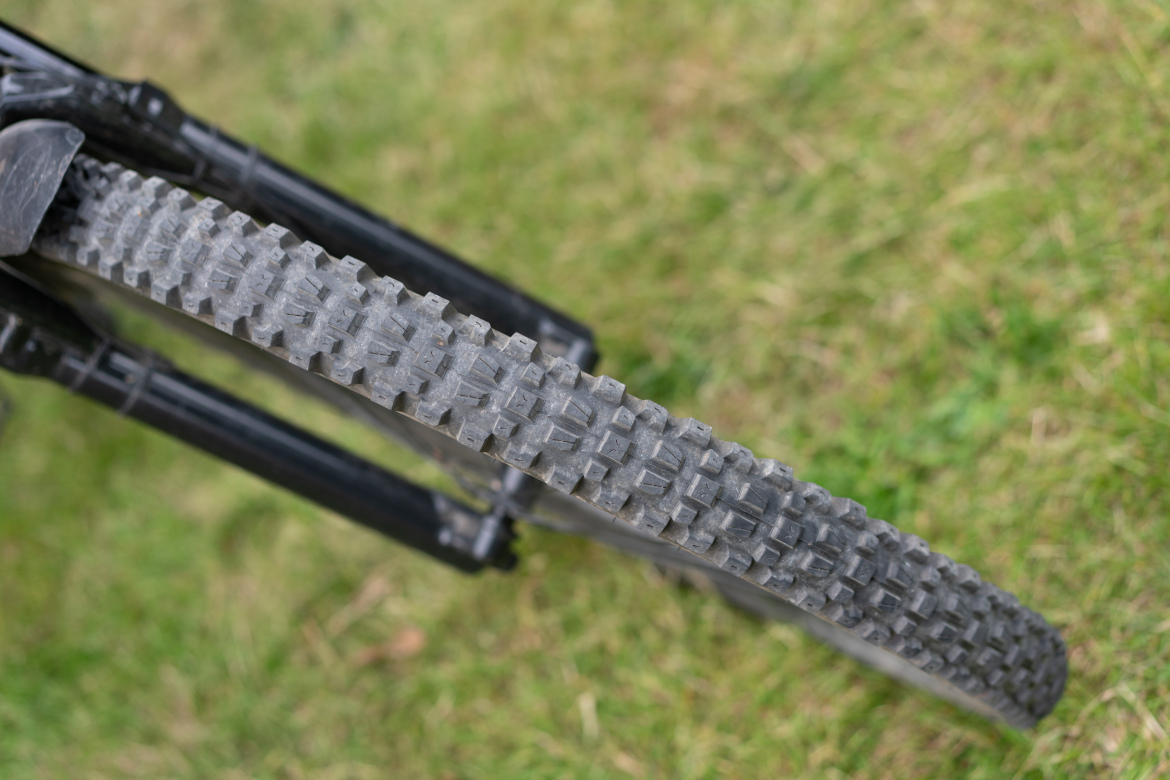 Close-up of a bicycle tire with a textured tread pattern, resting on green grass. The tire is shown from an angled perspective, highlighting the grip design suitable for various terrains.