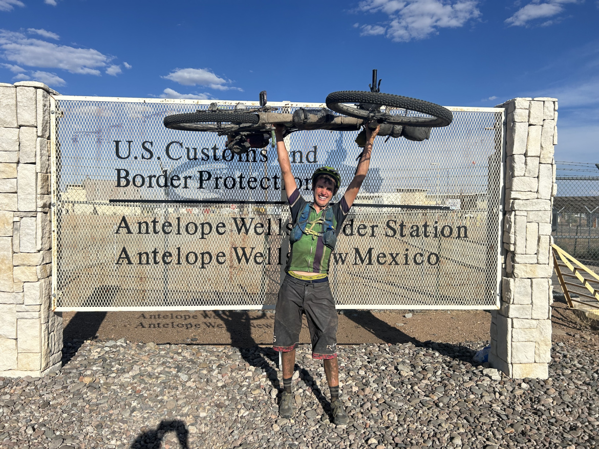 A cyclist stands in front of a U.S. Customs and Border Protection sign at Antelope Wells, New Mexico, holding their bike above their head in celebration. The background features a clear blue sky and rocky terrain.