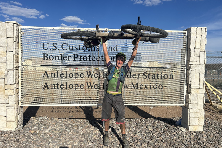 A cyclist stands in front of a U.S. Customs and Border Protection sign at Antelope Wells, New Mexico, holding their bike above their head in celebration. The background features a clear blue sky and rocky terrain.