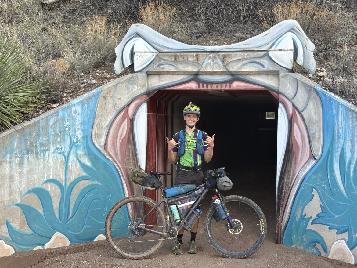 A smiling cyclist stands in front of a colorful mural on the entrance of a tunnel, holding up a hand gesture of approval. The cyclist is wearing a helmet and a brightly colored shirt, with a fully loaded bike beside them. The surrounding landscape features dry vegetation and earthy tones.